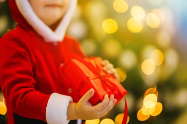 Bir çocuğun elinde Noel hediyesi. Noel Baba kostümlü küçük çocuğun elleri. Arka planda kırmızı hediye kutusu tutan komik bir çocuk. Tatiller, yeni yıl konsepti.