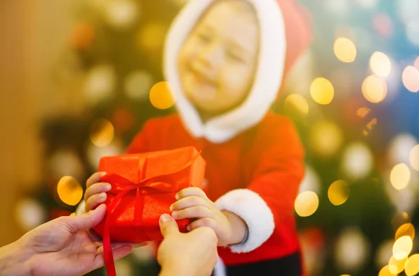 Hands of parent giving a hristmas gift to child. Happy child holding red gift box on the background lights. Kid in santa costume holds christmas present in hands. Holidays, presents, New year concept