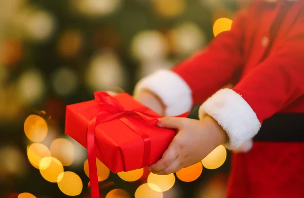 Christmas gift in the hands of a child. Hands of little boy in santa costume with Christmas present. Smiling funny kid holding red gift box on the background of lights. Holidays, New year concept.