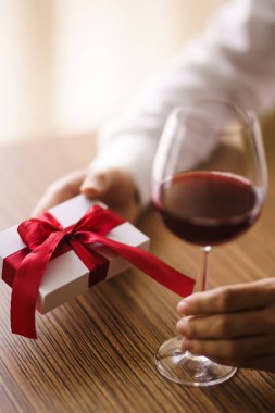 Bir bardak şarap ve hediyeyle bir adamın elleri. Kırmızı kurdeleli bir hediye. Erkek elleri sürpriz hediye kutusunu tutuyor. Sevgililer birbirlerine hediyeler verir. Sevgililer Günü konsepti. Romantik bir gün. Doğum günü yıldönümü.