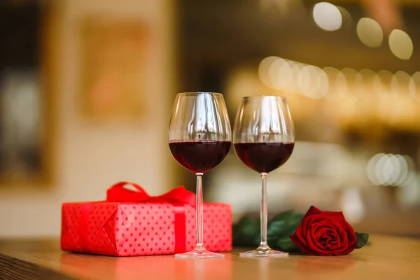 Glasses with wine, rose and red gift box on wooden table. Lovely romantic dinner. Romance at restaurant. Valentines day celebration concept. Relationship, surprise, Birthday concept.