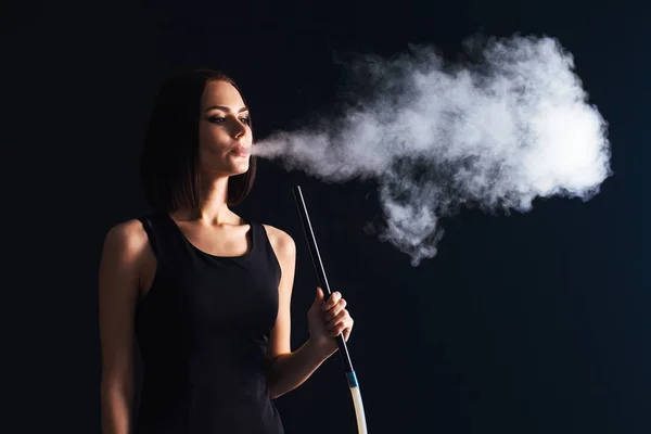 Young woman smoke a hookah on black wall background. The pleasure of smoking. Sexy smoke with vape. Exhaling smoke.
