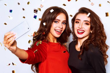 İki genç partinin fotoğrafında gülümseyen ve beyaz arka planda cep telefonuyla selfie çeken iki kadın. Instagram için konfeti partisinde selfie çeken güzel kızlar. Tatiller ve kutlama konsepti