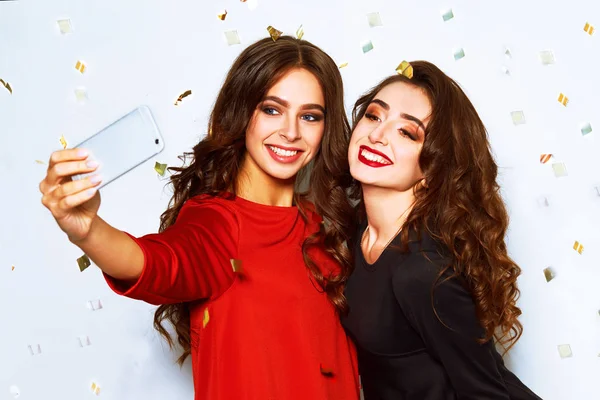 Photo of two young party women smiling and  making selfie photo on mobile phone over white background. Beautiful girls who make selfie for instagram on confetti party. Holidays and celebration concept