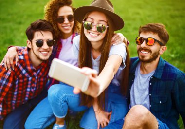 Bir grup güler yüzlü arkadaş çimlerin üzerinde oturup, yaz parkında selfie çekiyorlar. Gençler birlikte eğleniyor. Dinlenme, eğlence, yaz, teknoloji ve insan kavramı.