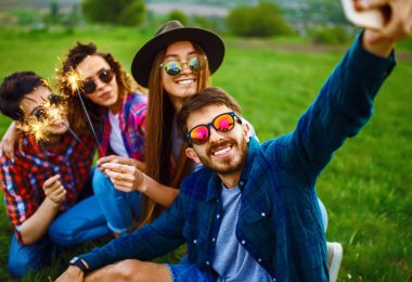 Bir grup güler yüzlü arkadaş çimlerin üzerinde oturup, yaz parkında selfie çekiyorlar. Gençler birlikte eğleniyor. Dinlenme, eğlence, yaz, teknoloji ve insan kavramı.