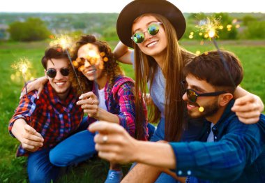 Bir grup arkadaş, yaz parkında çimenlerde oturan maytaplarla eğleniyor. Gençler birlikte eğleniyor. Bir grup genç eğleniyor. Dinlenme, eğlence, yaz, tatil konsepti.