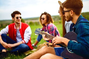 Gitarlı mutlu arkadaşlar parkta çimlerin üzerinde oturarak vakit geçiriyorlar. Adam gitar çalıyor. Gençler yaz parkında eğleniyor. Dinlenme, eğlence, yaz konsepti.