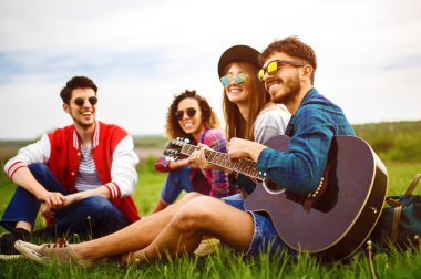 Gitarlı mutlu arkadaşlar parkta çimlerin üzerinde oturarak vakit geçiriyorlar. Adam gitar çalıyor. Gençler yaz parkında eğleniyor. Dinlenme, eğlence, yaz konsepti.