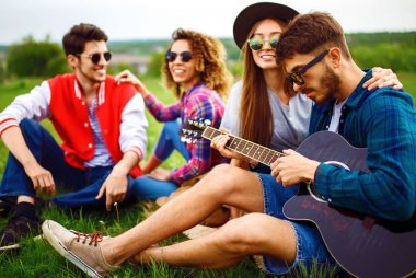 Gitarlı mutlu arkadaşlar parkta çimlerin üzerinde oturarak vakit geçiriyorlar. Adam gitar çalıyor. Gençler yaz parkında eğleniyor. Dinlenme, eğlence, yaz konsepti.