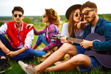 Gitarlı mutlu arkadaşlar parkta çimlerin üzerinde oturarak vakit geçiriyorlar. Adam gitar çalıyor. Gençler yaz parkında eğleniyor. Dinlenme, eğlence, yaz konsepti.