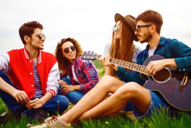 Gitarlı mutlu arkadaşlar parkta çimlerin üzerinde oturarak vakit geçiriyorlar. Adam gitar çalıyor. Gençler yaz parkında eğleniyor. Dinlenme, eğlence, yaz konsepti.
