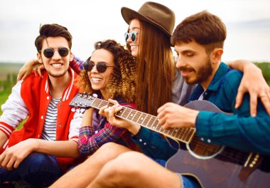Gitarlı mutlu arkadaşlar parkta çimlerin üzerinde oturarak vakit geçiriyorlar. Adam gitar çalıyor. Gençler yaz parkında eğleniyor. Dinlenme, eğlence, yaz konsepti.