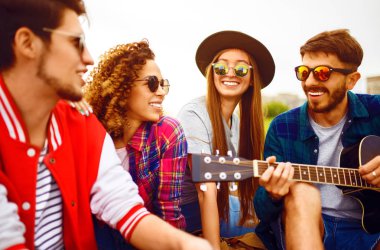 Gitarlı mutlu arkadaşlar parkta çimlerin üzerinde oturarak vakit geçiriyorlar. Adam gitar çalıyor. Gençler yaz parkında eğleniyor. Dinlenme, eğlence, yaz konsepti.