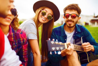 Gitarlı mutlu arkadaşlar parkta çimlerin üzerinde oturarak vakit geçiriyorlar. Adam gitar çalıyor. Gençler yaz parkında eğleniyor. Dinlenme, eğlence, yaz konsepti.