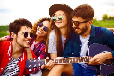 Gitarlı mutlu arkadaşlar parkta çimlerin üzerinde oturarak vakit geçiriyorlar. Adam gitar çalıyor. Gençler yaz parkında eğleniyor. Dinlenme, eğlence, yaz konsepti.