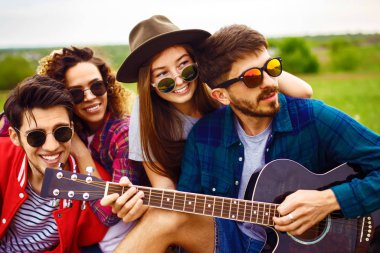 Gitarlı mutlu arkadaşlar parkta çimlerin üzerinde oturarak vakit geçiriyorlar. Adam gitar çalıyor. Gençler yaz parkında eğleniyor. Dinlenme, eğlence, yaz konsepti.
