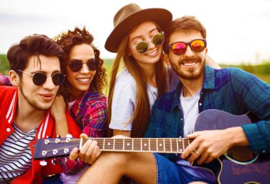 Gitarlı mutlu arkadaşlar parkta çimlerin üzerinde oturarak vakit geçiriyorlar. Adam gitar çalıyor. Gençler yaz parkında eğleniyor. Dinlenme, eğlence, yaz konsepti.