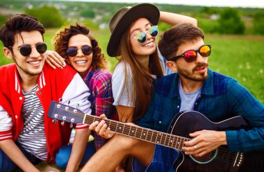 Gitarlı mutlu arkadaşlar parkta çimlerin üzerinde oturarak vakit geçiriyorlar. Adam gitar çalıyor. Gençler yaz parkında eğleniyor. Dinlenme, eğlence, yaz konsepti.