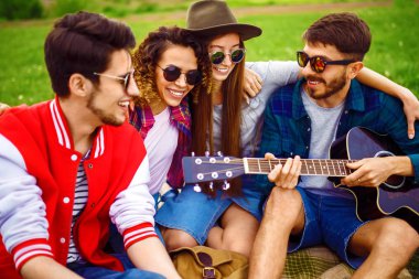 Gitarlı mutlu arkadaşlar parkta çimlerin üzerinde oturarak vakit geçiriyorlar. Adam gitar çalıyor. Gençler yaz parkında eğleniyor. Dinlenme, eğlence, yaz konsepti.
