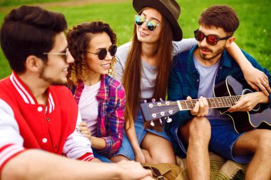 Gitarlı mutlu arkadaşlar parkta çimlerin üzerinde oturarak vakit geçiriyorlar. Adam gitar çalıyor. Gençler yaz parkında eğleniyor. Dinlenme, eğlence, yaz konsepti.