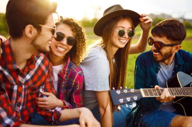 Gitarlı mutlu arkadaşlar parkta çimlerin üzerinde oturarak vakit geçiriyorlar. Adam gitar çalıyor. Gençler yaz parkında eğleniyor. Dinlenme, eğlence, yaz konsepti.