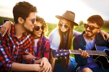 Gitarlı mutlu arkadaşlar parkta çimlerin üzerinde oturarak vakit geçiriyorlar. Adam gitar çalıyor. Gençler yaz parkında eğleniyor. Dinlenme, eğlence, yaz konsepti.