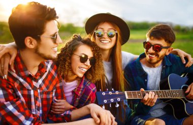 Gitarlı mutlu arkadaşlar parkta çimlerin üzerinde oturarak vakit geçiriyorlar. Adam gitar çalıyor. Gençler yaz parkında eğleniyor. Dinlenme, eğlence, yaz konsepti.
