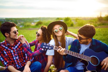 Gitarlı mutlu arkadaşlar parkta çimlerin üzerinde oturarak vakit geçiriyorlar. Adam gitar çalıyor. Gençler yaz parkında eğleniyor. Dinlenme, eğlence, yaz konsepti.