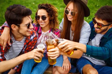 Mutlu arkadaşlar parkta birlikte vakit geçiriyorlar çimenlerde oturup, bira içip sohbet ediyorlar. Bir grup genç yaz parkında eğleniyor. Dinlenme, eğlence, yaz konsepti.