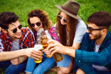 Mutlu arkadaşlar parkta birlikte vakit geçiriyorlar çimenlerde oturup, bira içip sohbet ediyorlar. Bir grup genç yaz parkında eğleniyor. Dinlenme, eğlence, yaz konsepti.