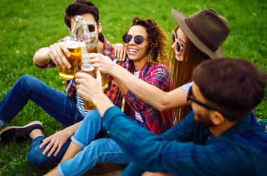 Mutlu arkadaşlar parkta birlikte vakit geçiriyorlar çimenlerde oturup, bira içip sohbet ediyorlar. Bir grup genç yaz parkında eğleniyor. Dinlenme, eğlence, yaz konsepti.