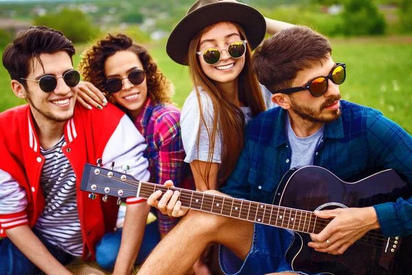Gitarlı mutlu arkadaşlar parkta çimlerin üzerinde oturarak vakit geçiriyorlar. Adam gitar çalıyor. Gençler yaz parkında eğleniyor. Dinlenme, eğlence, yaz konsepti.
