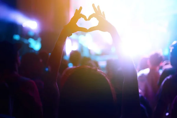 Heart shaped hands at concert, loving the artist and the festival. Audience with hands raised at a music festival and lights streaming down from above the stage.