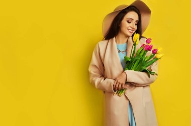 Güzel bir ceket ve şapka giymiş, sarı arka planda çiçek laleleri olan güzel bir kız. Genç bir kadın hediye olarak çiçek aldı ve sevindi. Çiçekli bahar kızı. 8 Mart. 