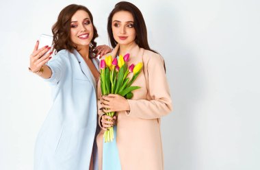 İki güzel kız ellerinde lalelerle selfie çekiyorlar. Moda kıyafetleri içinde poz verip gülümseyen iki genç kadın. Çiçekli bahar kızı. 8 Mart.