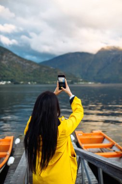 Kız turist, Norveç 'teki gölün kenarında telefonla fotoğraf çekiyor. Norveç 'teki dağların arka planına karşı gölün kenarında dinlenen aktif bir kadın. Seyahat, yaşam tarzı, macera konsepti.