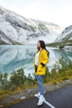 Norveç 'teki gölde poz veren sarı ceketli kız. Norveç 'teki dağların arka planına karşı gölün kenarında dinlenen aktif bir kadın. Seyahat, yaşam tarzı, macera, vahşi doğa.