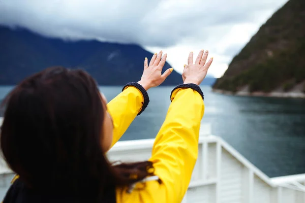 Young woman enjoys freedom against the backdrop of the mountains in the Norway. The girl tourist in a yellow jacket looking on the majestic mountains and pulls her hands to the sky.