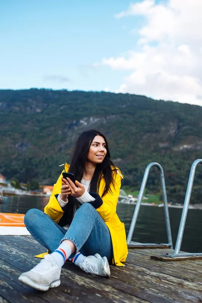 The girl tourist takes a photo on the phone by the lake in Norway. Young woman takes selfie against the backdrop of the mountains in the Norway. Travelling, lifestyle, adventure, concept.