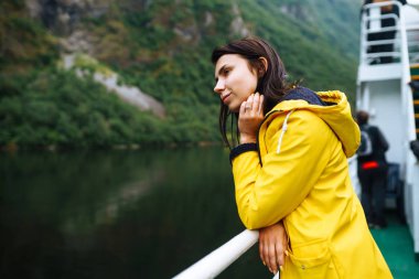 The girl tourist on a pleasure boat on the fjord enjoys the picturesque mountains and lakes of Norway. Young woman posing against the backdrop of the mountains. Travelling, lifestyle, adventure.