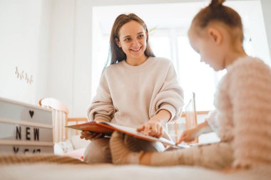 Neşeli genç anne küçük kızına kitap okuyor. Sevimli bir anne ve kız, beyaz bir odada uzanmış kitap okuyor ve yatakta dinleniyor. Mutlu sevgi dolu aile. 