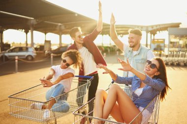 Genç arkadaşlar açık havada alışveriş arabalarında eğleniyorlar. Bir grup mutlu genç yazın güneş ışığıyla alışveriş arabasında yarışıyor. Yaşam tarzı, eğlence, eğlence, gençlik kavramı.       