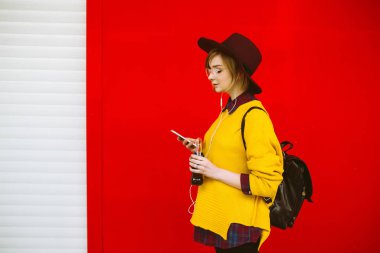 İçkili ve telefonlu genç bir kız. Renkli arka planda poz veriyor. Sarı kazaklı ve şapkalı hippi kız camdan içiyor ve cep telefonuyla mesajlaşıyor. Yaşam tarzı, gençlik kavramı.