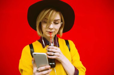 İçkili ve telefonlu genç bir kız. Renkli arka planda poz veriyor. Sarı kazaklı ve şapkalı hippi kız camdan içiyor ve cep telefonuyla mesajlaşıyor. Yaşam tarzı, gençlik kavramı.