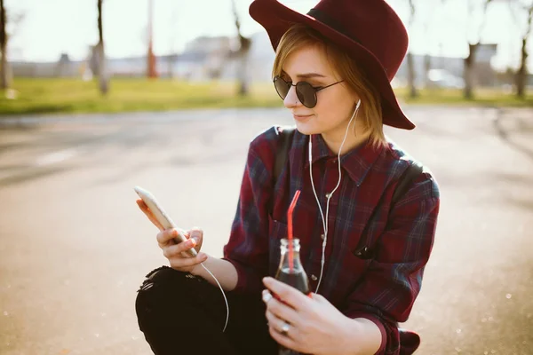 stock image Young teenager girl sitting at the skatepark, holding mobile phone while listening to music with headphones. Pretty woman  in sunglasses and hat drinking from glass, with phone sitting on skateboard. 