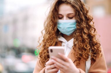 Yüzünde karantina şehrinde bir telefonla steril tıbbi maske takan bir kız. Haber aramak için telefonu kullanan bir kadın. Salgının yayılmasını önleme konsepti.