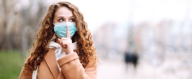 Uzun kenar. Yüzünde steril tıbbi maske takan kız bir jest gösteriyor: Sessizlik. Salgının yayılmasını önleme ve koronavirüsü tedavi etme kavramı karantina şehrinde salgındır..