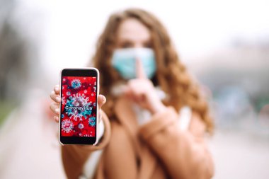 Yüzünde steril tıbbi maske takan kız telefonu karantina şehrinde tutuyor. Coronavirus hücreleri telefon ekranında. Salgının yayılmasını önleme konsepti. 