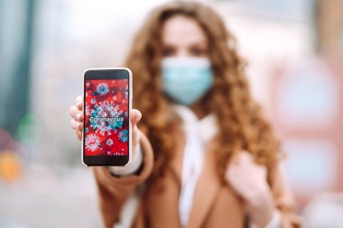Yüzünde steril tıbbi maske takan kız telefonu karantina şehrinde tutuyor. Coronavirus hücreleri telefon ekranında. Salgının yayılmasını önleme konsepti. 